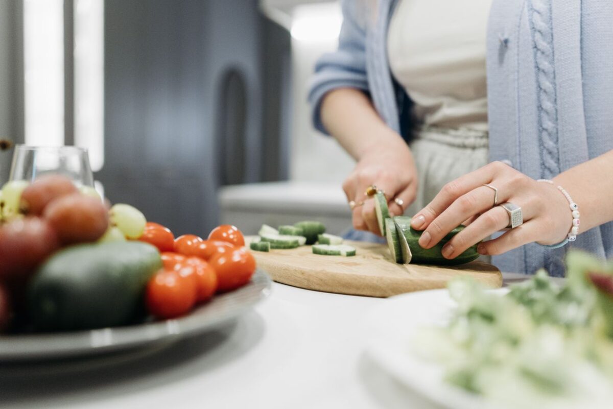 home-services-02 Close-up of a woman slicing cucumbers on a wooden board surrounded by fresh vegetables.